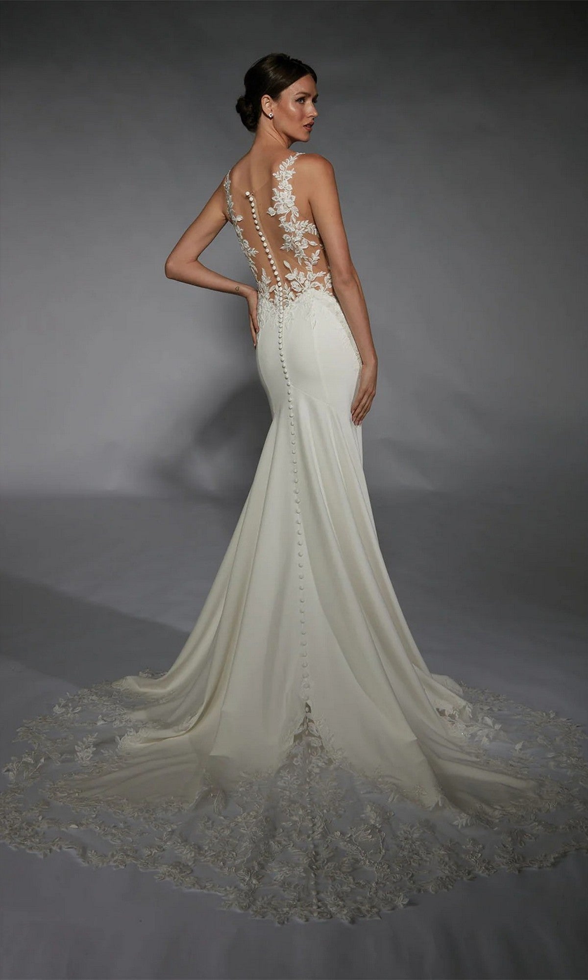 A woman poses in the Alyce 7117 long white dress, featuring a fitted silhouette, ivory hue, sheer back, floral lace accents, fabric-covered buttons, and a long lace train against a gray background.